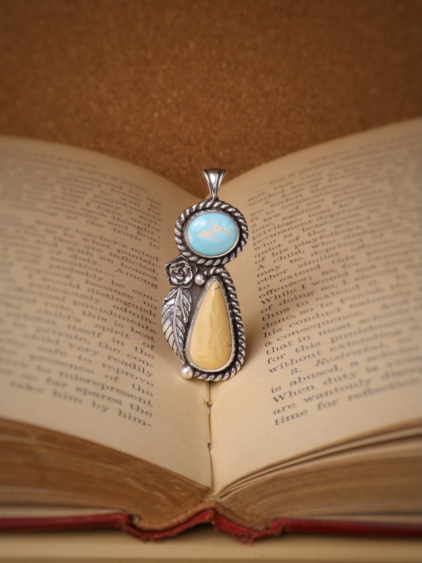 A silver-toned pendant with a turquoise oval stone and a teardrop-shaped cream-toned stone, featuring a small rose detail, resting on the pages of an open book,
