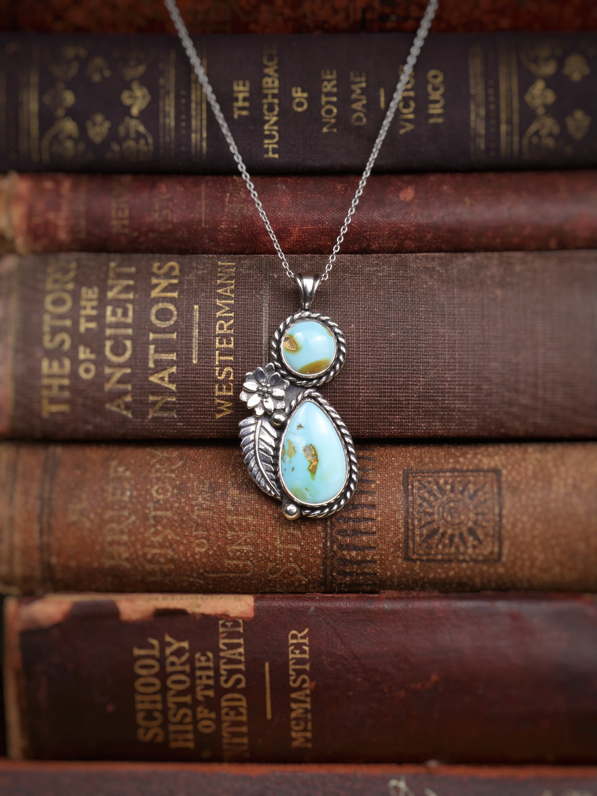 A silver pendant necklace with two turquoise stones, displayed on top of a stack of vintage brown leather-bound books.