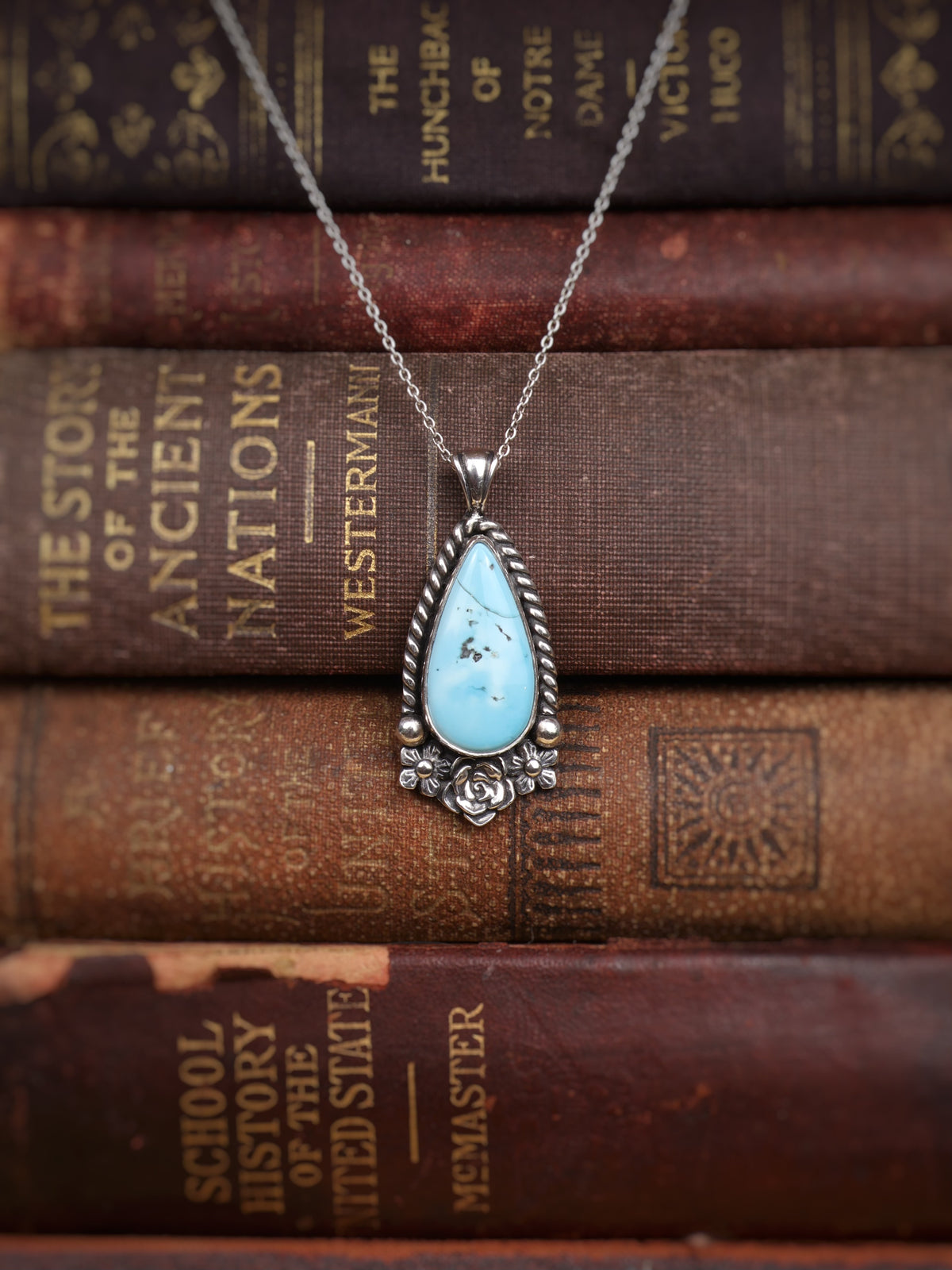 A silver teardrop pendant necklace with a turquoise stone and three small flowers, displayed on a stack of vintage brown books.