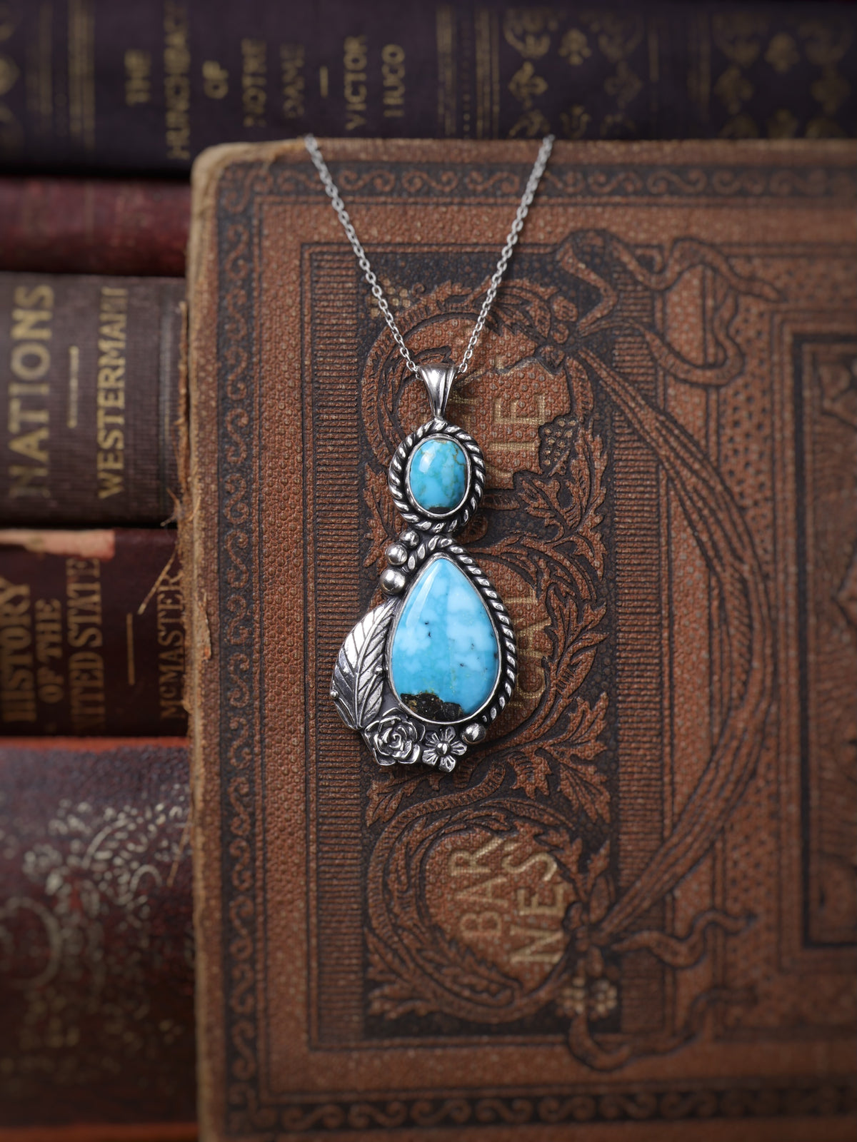 A silver pendant necklace with a large teardrop-shaped turquoise stone and a smaller round turquoise stone, displayed on an ornate brown vintage book with other books in the background.