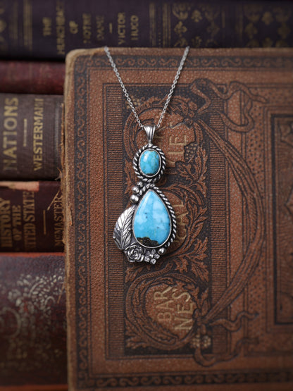 A silver pendant necklace with a large teardrop-shaped turquoise stone and a smaller round turquoise stone, displayed on an ornate brown vintage book with other books in the background.