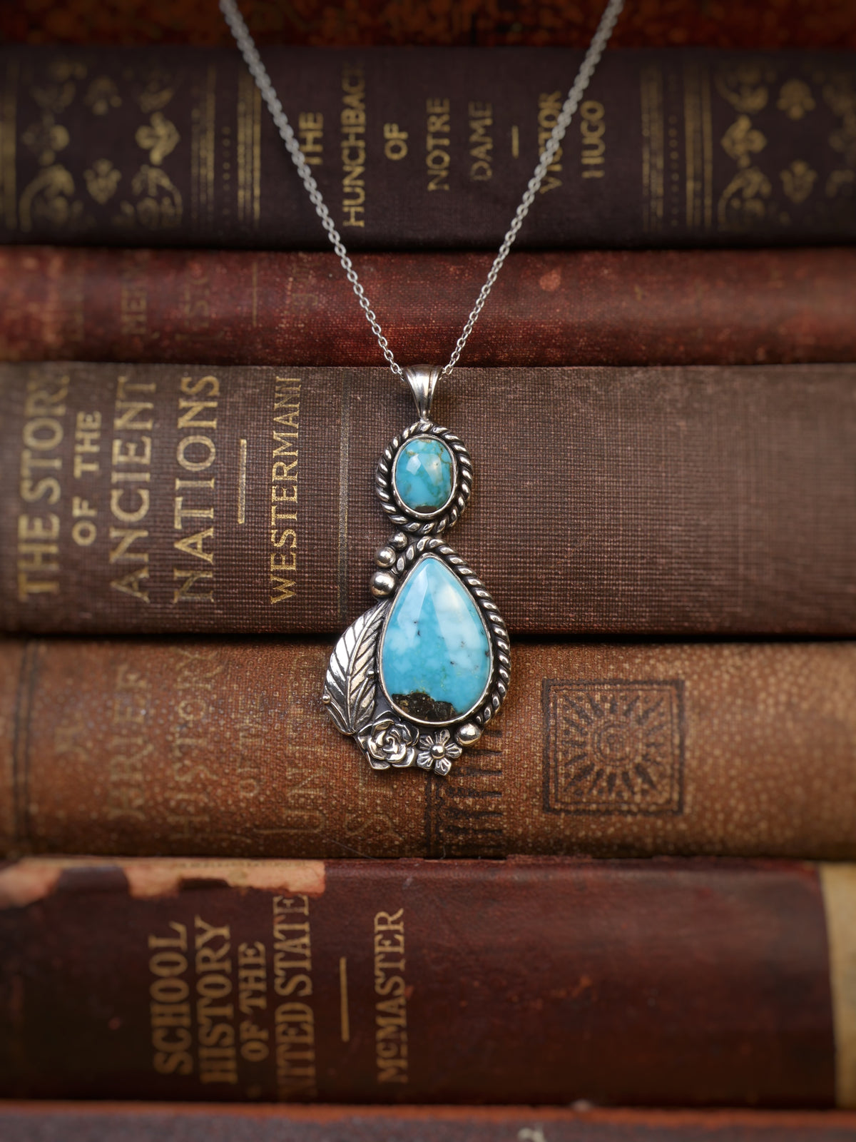 A silver pendant necklace with a teardrop-shaped turquoise stone and a smaller round turquoise stone, displayed on top of a stack of vintage brown book spines.