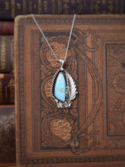 A silver pendant necklace featuring a turquoise stone, surrounded by a silver leaf and rose, displayed on an antique brown book.