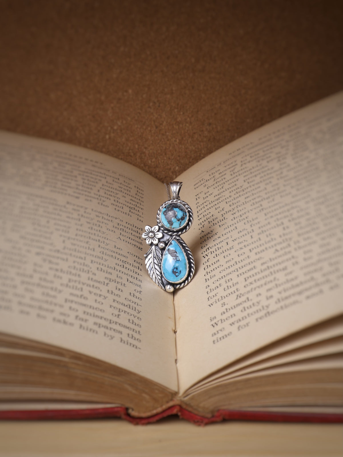 A silver pendant with two turquoise stones and a small flower detail, resting on the open pages of an antique book against a brown background.