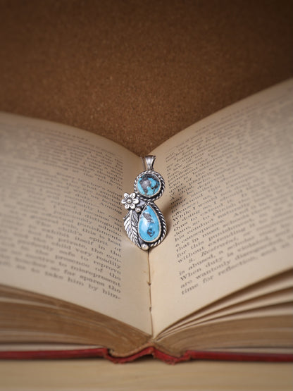 A silver pendant with two turquoise stones and a small flower detail, resting on the open pages of an antique book against a brown background.