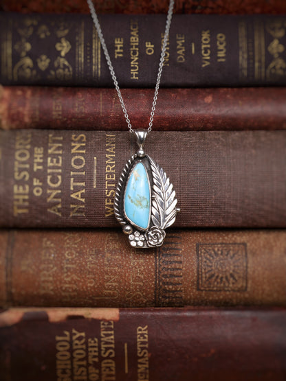 A silver pendant necklace with a turquoise stone, displayed on top of vintage brown leather-bound books.