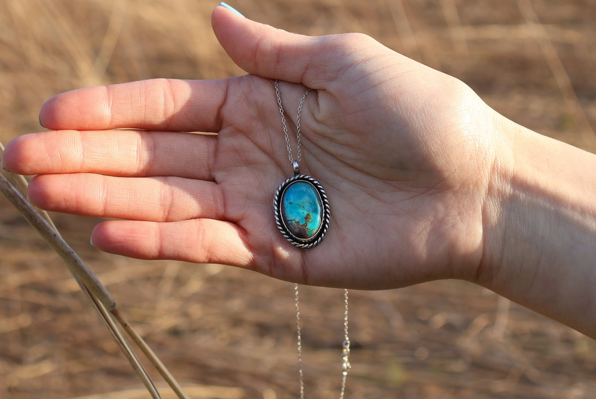 Hand holding a necklace with a turquoise pendant against a natural background