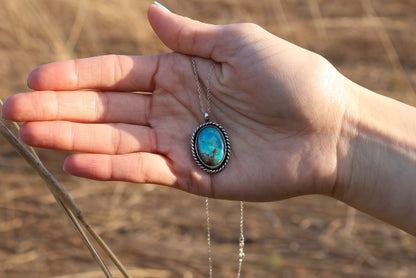 Hand holding a necklace with a turquoise pendant against a natural background