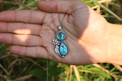 A hand holding a silver pendant necklace with two turquoise stones and a small silver flower detail, set against a blurred outdoor background of green grass.