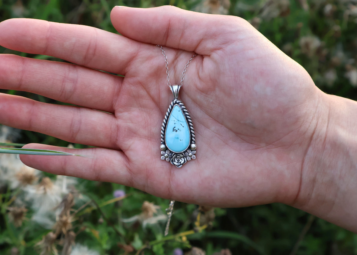 A silver teardrop pendant necklace with a turquoise stone, held in an open hand against a blurred green outdoor background.