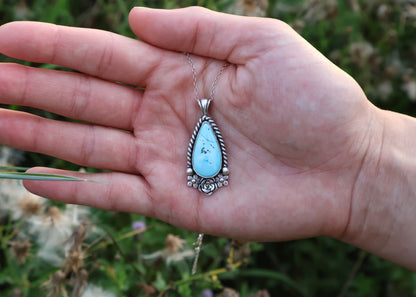 A silver teardrop pendant necklace with a turquoise stone, held in an open hand against a blurred green outdoor background.