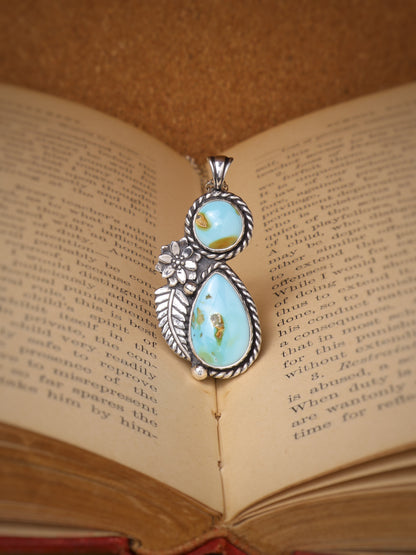 A silver pendant with two light blue turquoise stones and a small flower detail, resting on the pages of an open book.