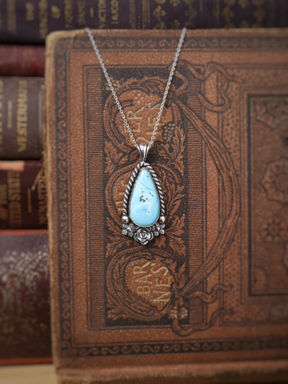 A silver teardrop pendant necklace with a turquoise stone, displayed on an ornate vintage brown book with other books blurred in the background.
