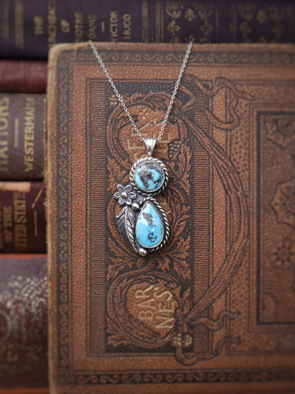 A silver pendant necklace with two turquoise stones and a small floral detail, displayed on an ornate brown book cover with other vintage books in the background.