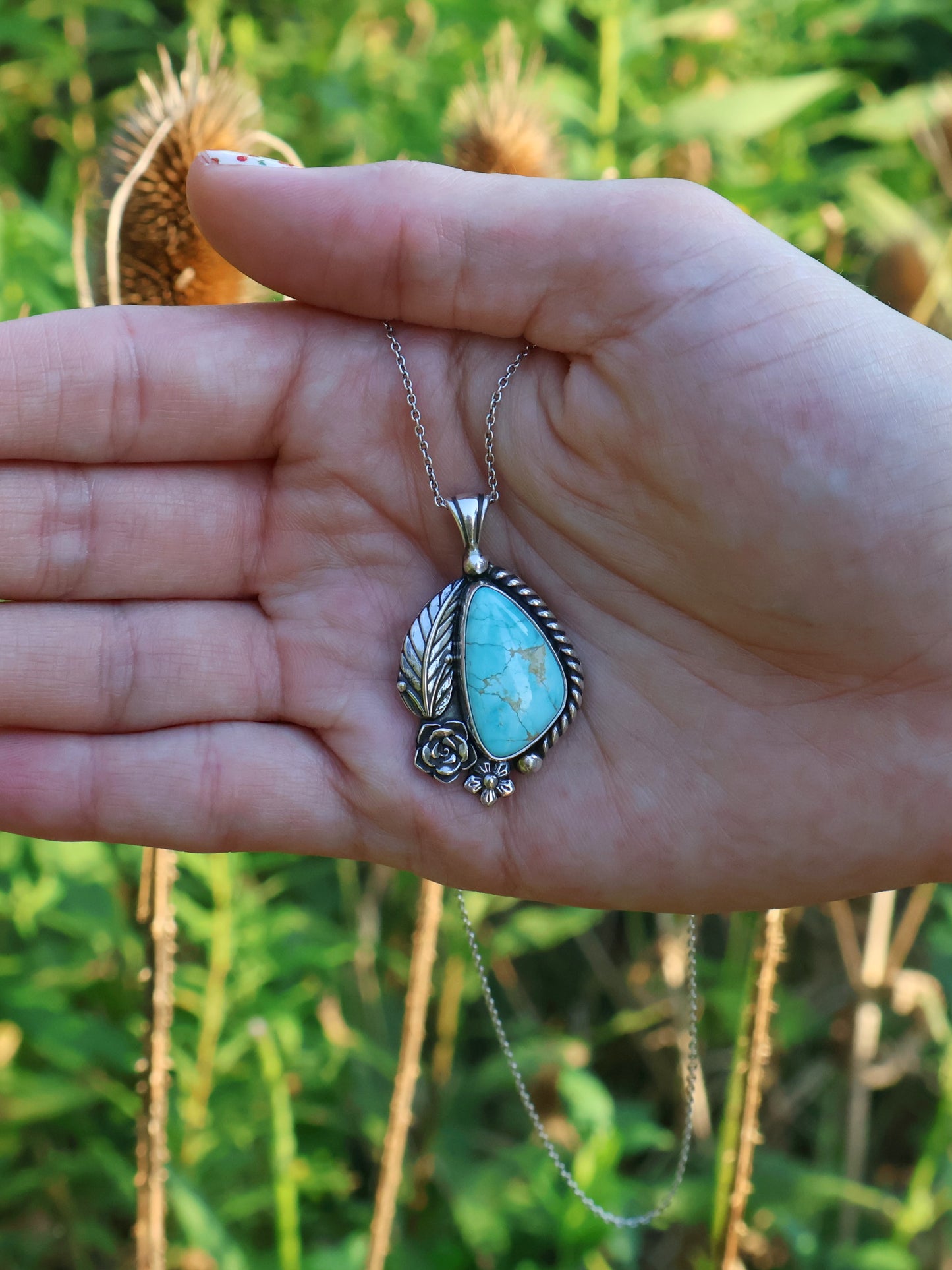 A silver pendant with a turquoise stone surrounded by a silver leaf, rose, and flower. A hand is holding it in front of a grassy background. 