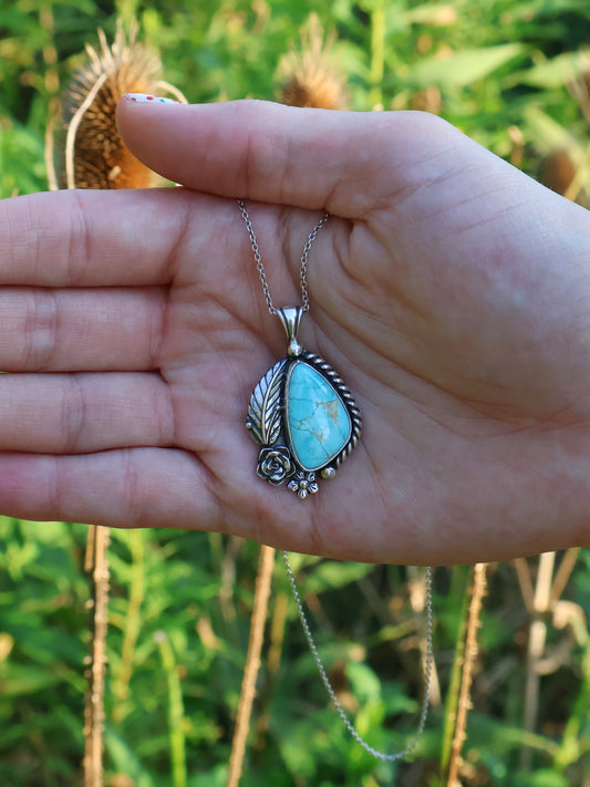 A silver pendant with a turquoise stone surrounded by a silver leaf, rose, and flower. A hand is holding it in front of a grassy background. 