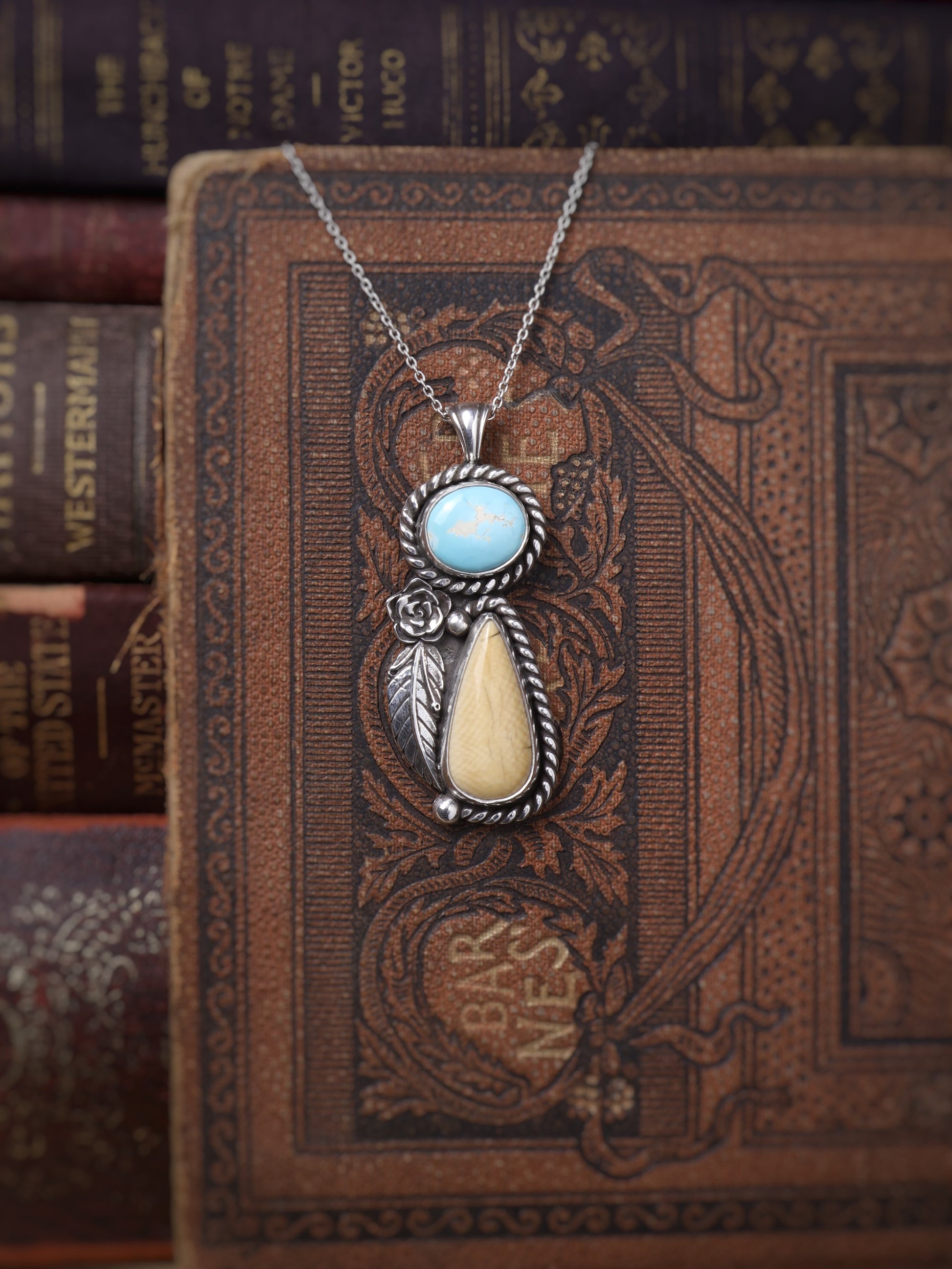 A silver pendant necklace with a turquoise oval stone and a cream teardrop stone, displayed on an antique brown book with other books blurred in the background.