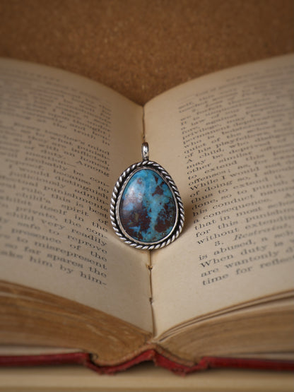 A silver oval pendant with a turquoise-blue marbled stone and a twisted silver border, resting on the edge of an open vintage book.