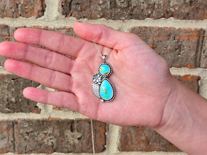 A sterling silver pendant necklace with two turquoise stones is held in a person's hand against a rustic brick wall background.