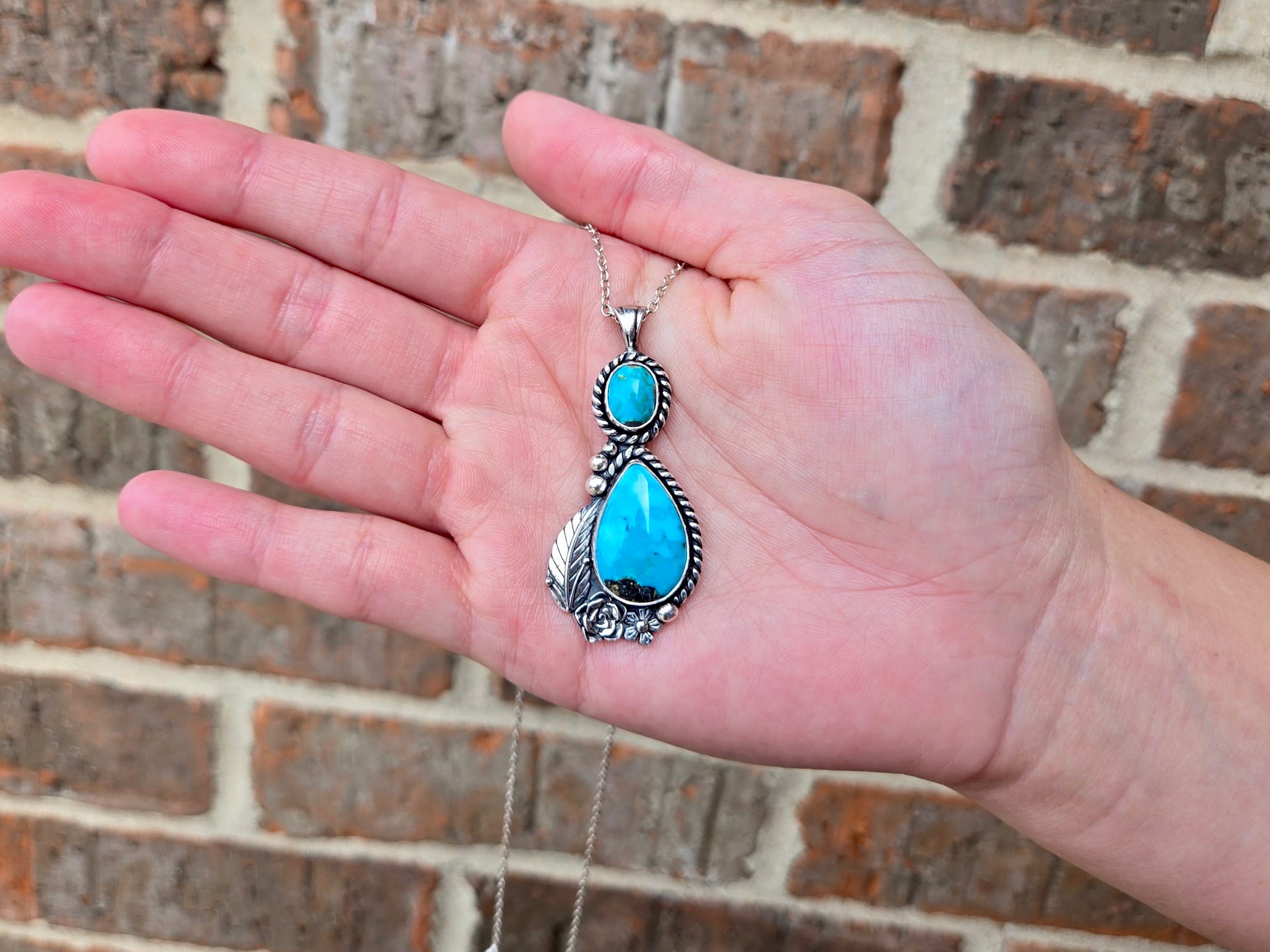 A hand holding a silver pendant necklace with a large turquoise teardrop stone and a smaller round turquoise stone, set against a brick wall background.