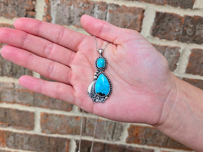 A hand holding a silver pendant necklace with a large turquoise teardrop stone and a smaller round turquoise stone, set against a brick wall background.
