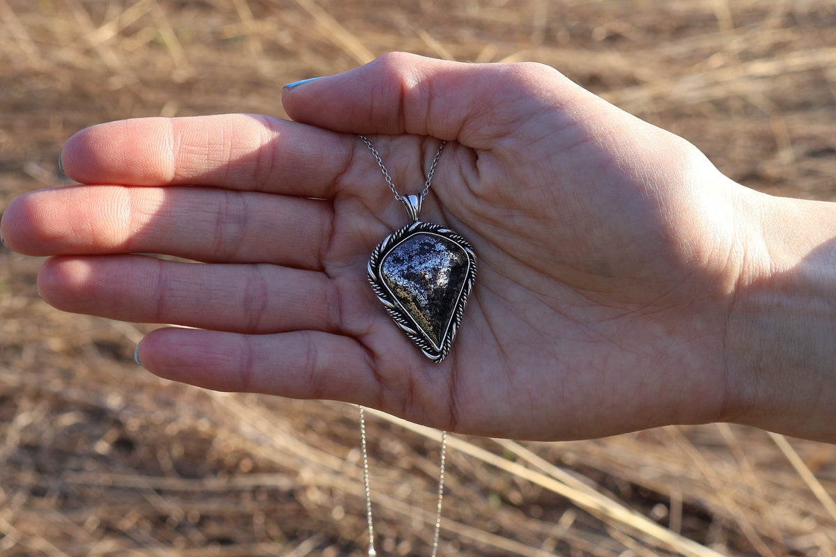 Hand holding a necklace with a silver pendant against a natural background