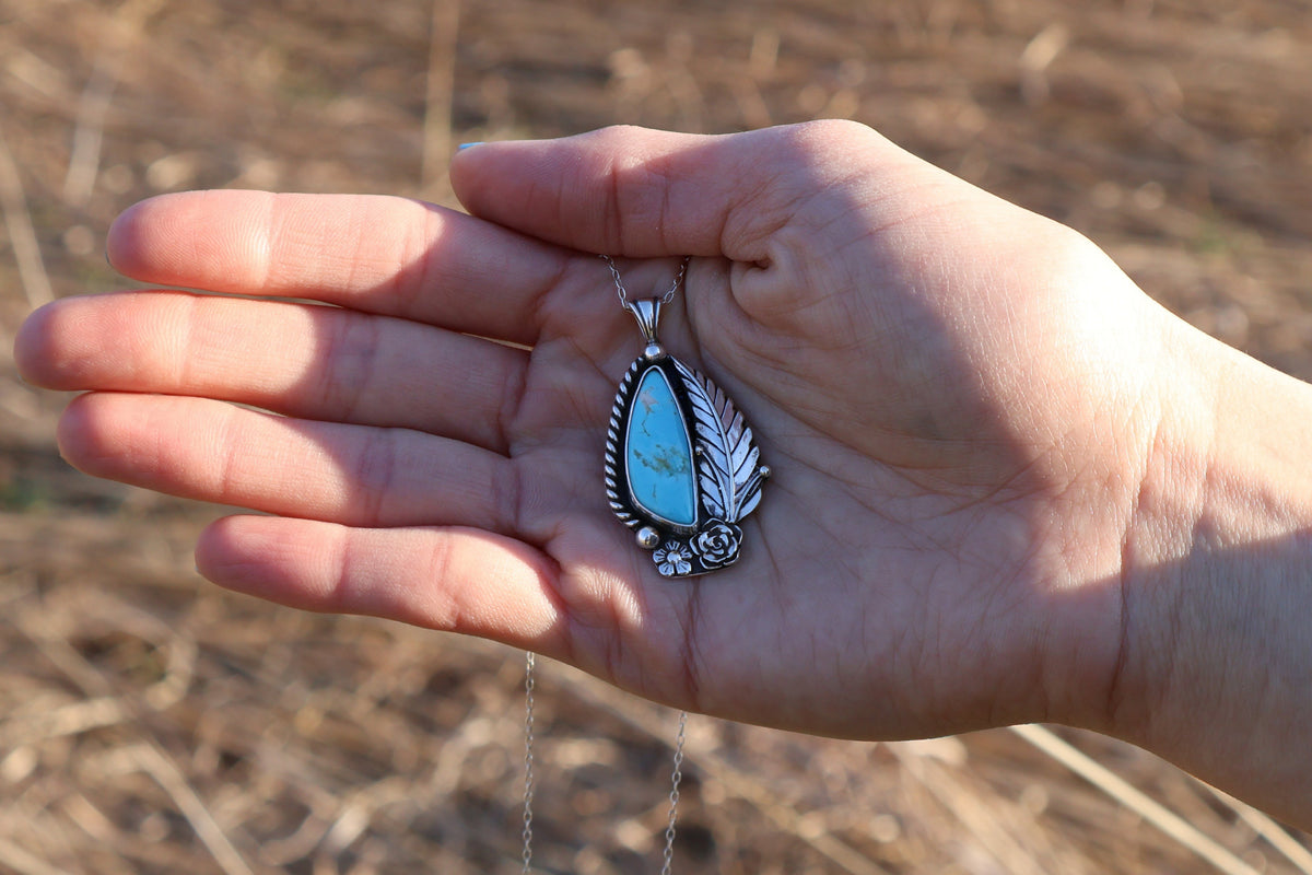 Hand holding a silver pendant with turquoise stone against a blurred natural background