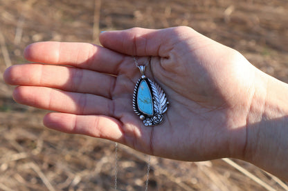 Hand holding a silver pendant with turquoise stone against a blurred natural background