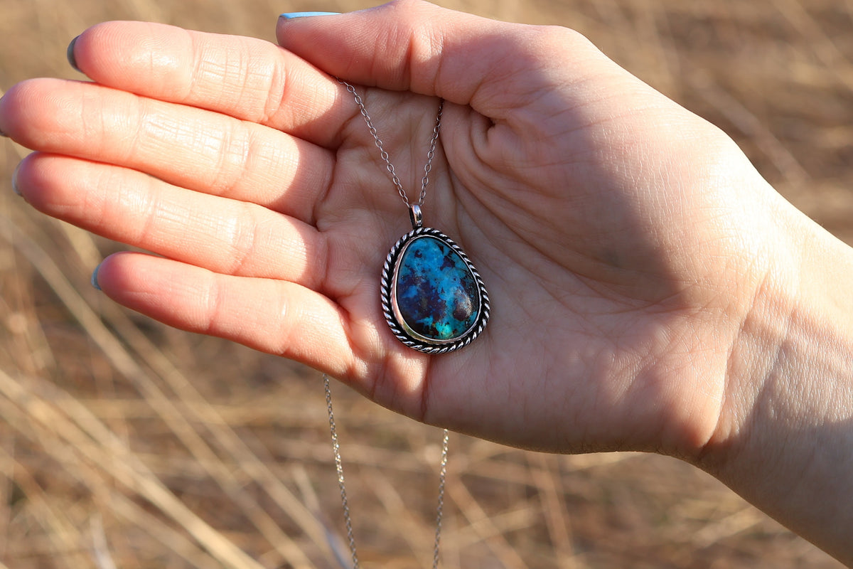 Hand holding a necklace with a turquoise pendant against a natural background