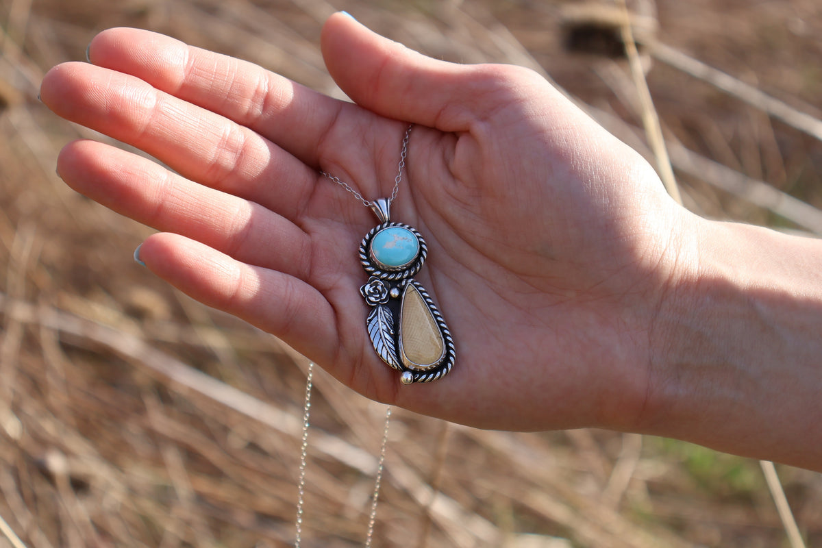 Hand holding a necklace with a cream toned stone and turquoise stone pendant against a natural background