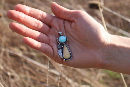 Hand holding a necklace with a cream toned stone and turquoise stone pendant against a natural background
