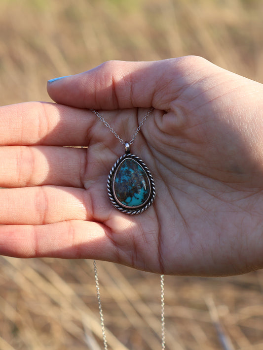 A silver necklace with a teardrop-shaped turquoise stone pendant, held in a person's hand against a blurred outdoor background of dry grass.