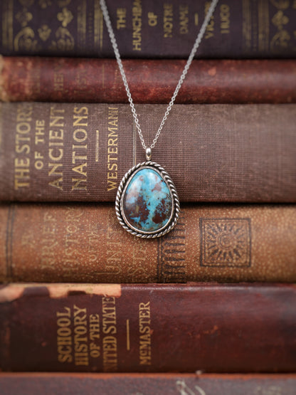 A silver pendant necklace with a turquoise stone, displayed on a stack of vintage brown leather-bound books.