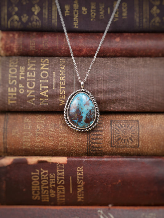 A silver pendant necklace with a turquoise stone, displayed on a stack of vintage brown leather-bound books.