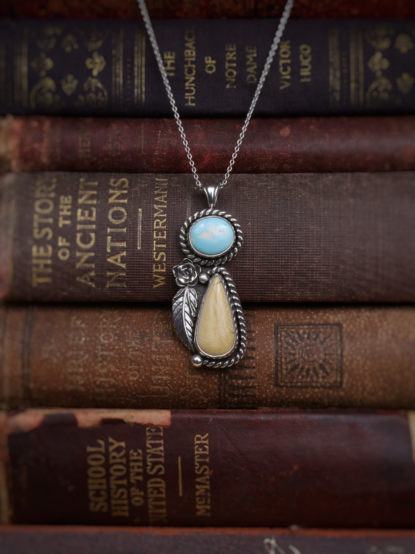 A sterling silver pendant necklace with a turquoise stone and a teardrop-shaped cream stone, displayed on a stack of vintage book spines.