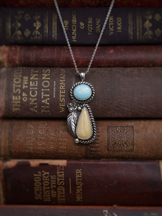 A sterling silver pendant necklace with a turquoise stone and a teardrop-shaped cream stone, displayed on a stack of vintage book spines.