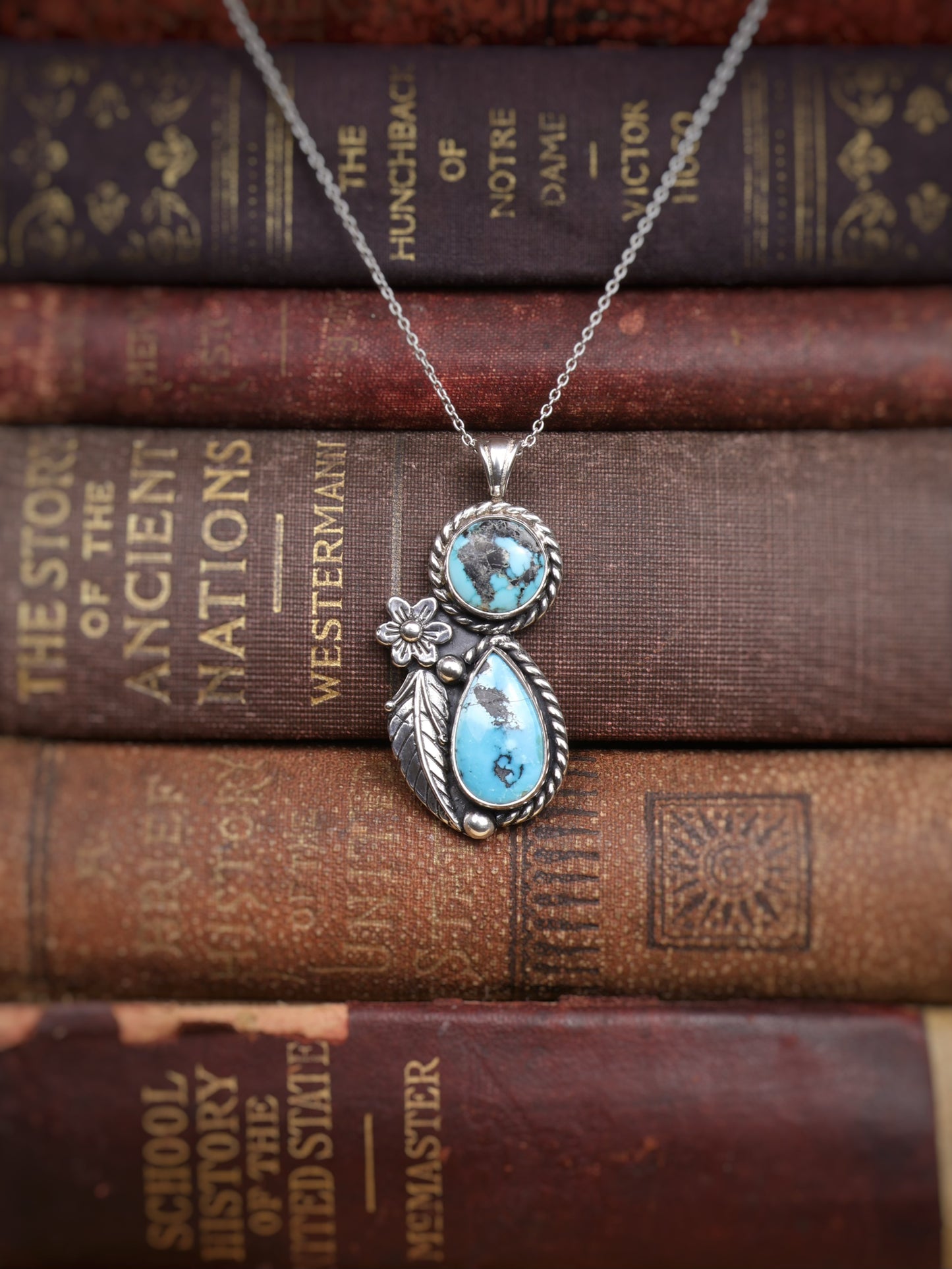 A silver pendant necklace with two turquoise stones and a small flower detail, displayed on top of stacked vintage book spines.