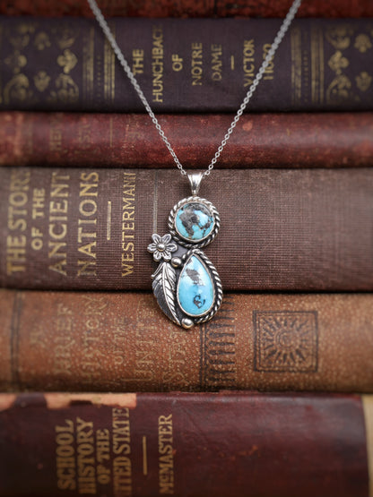 A silver pendant necklace with two turquoise stones and a small flower detail, displayed on top of stacked vintage book spines.