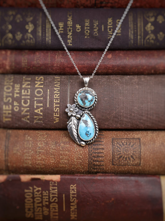 A silver pendant necklace with two turquoise stones and a small flower detail, displayed on top of stacked vintage book spines.