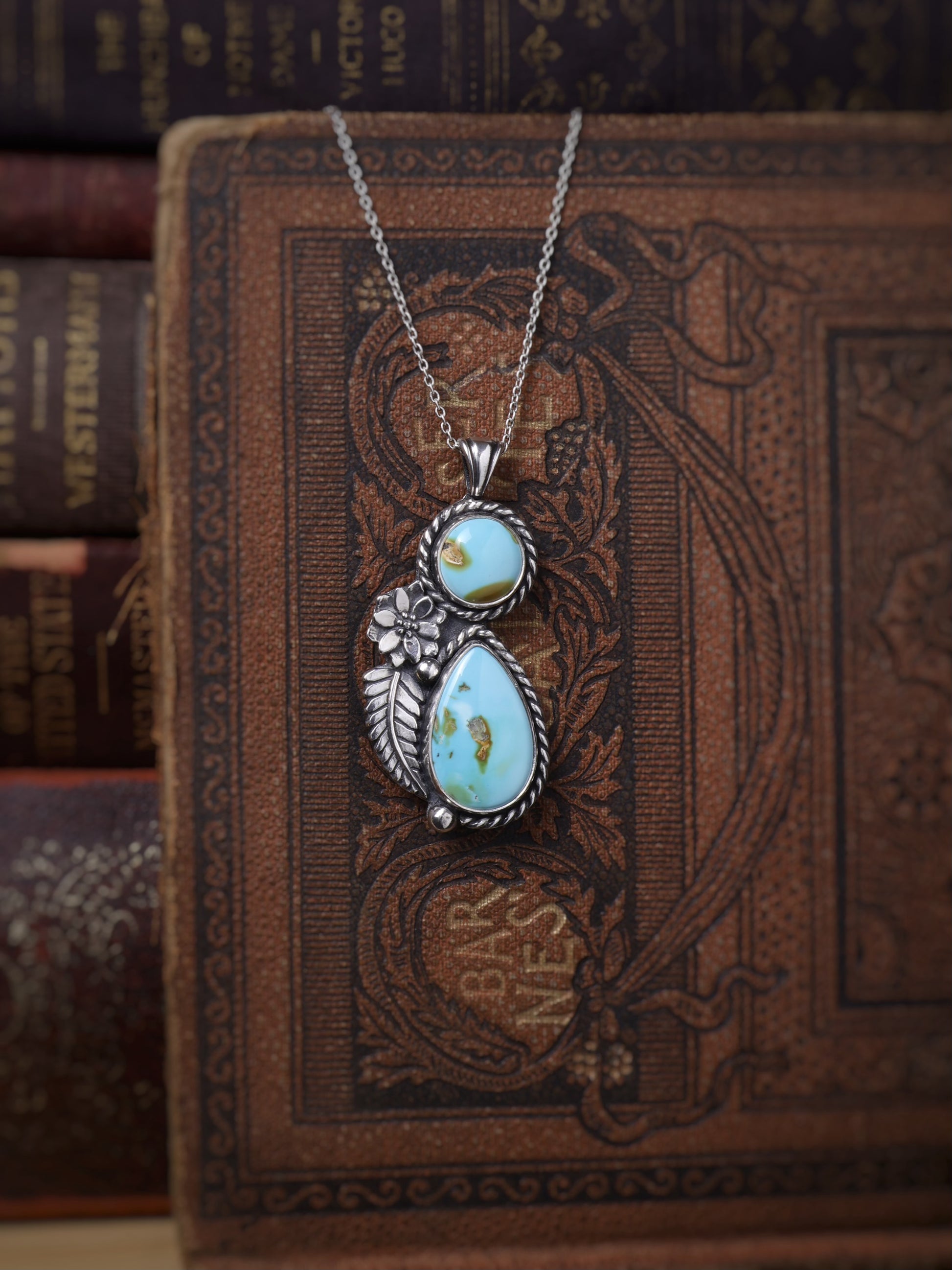 A silver pendant necklace with two light blue turquoise stones and a small floral detail, displayed on an ornate vintage brown book.