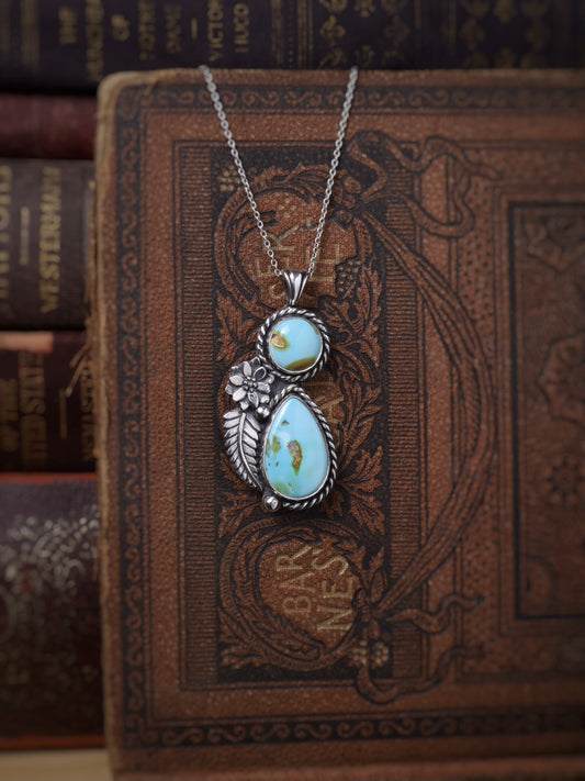 A silver pendant necklace with two light blue turquoise stones and a small floral detail, displayed on an ornate vintage brown book.