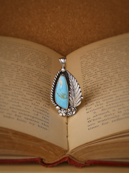 A silver pendant with a turquoise stone, displayed on the pages of an open vintage book.