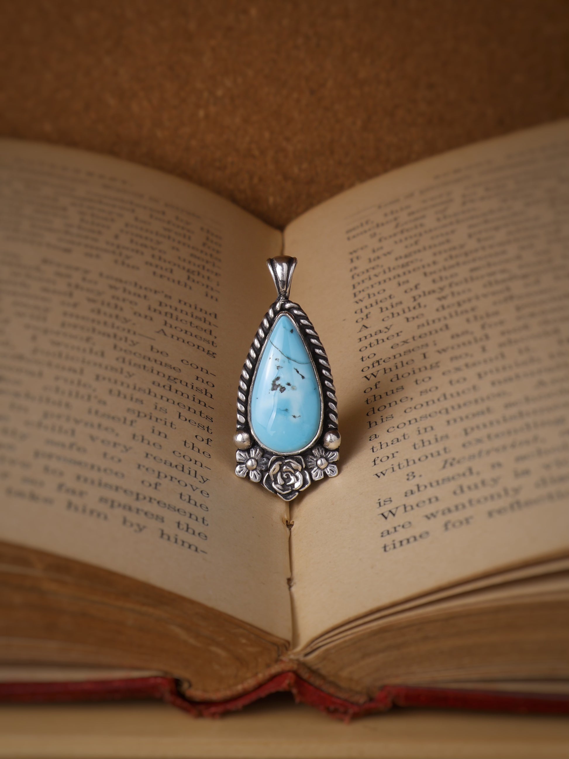 A silver teardrop pendant with a light blue turquoise center stone with a small rose and two flowers at the base, resting on the pages of an open vintage book.