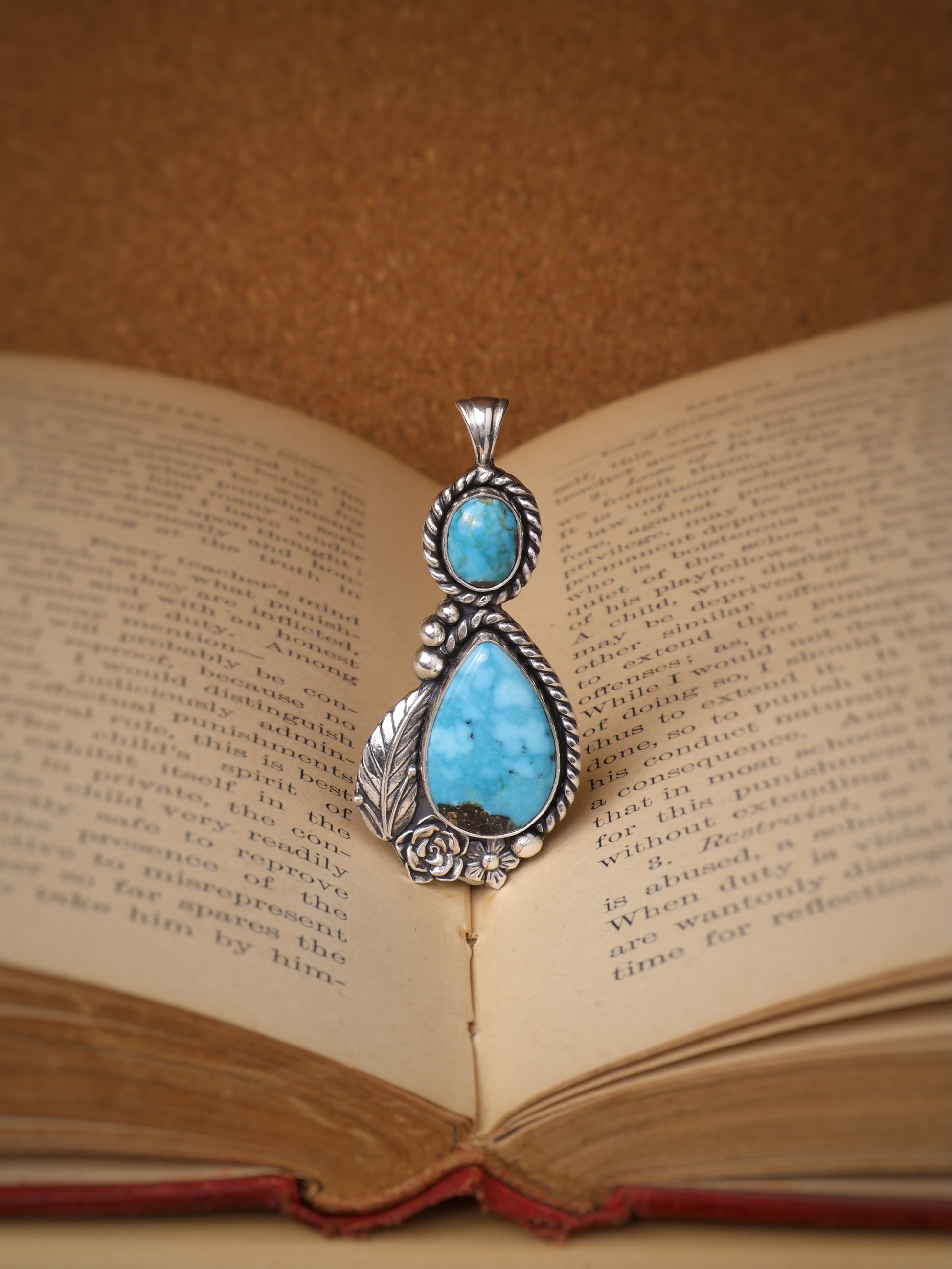 A silver pendant with a large turquoise stone and a smaller round turquoise stone, displayed on the pages of an open vintage book against a warm brown background.