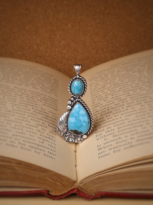 A silver pendant with a large turquoise stone and a smaller round turquoise stone, displayed on the pages of an open vintage book against a warm brown background.