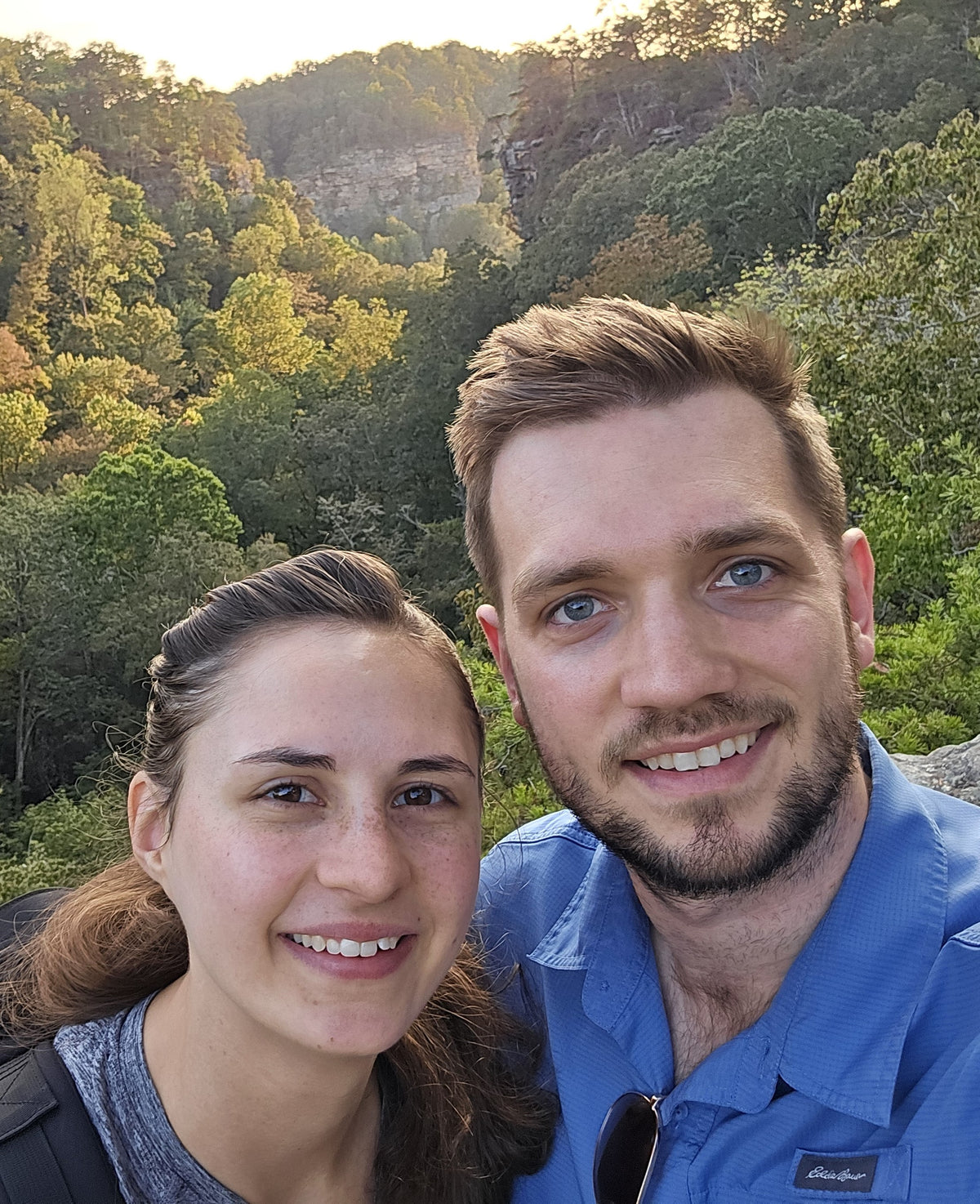Two people posing for a selfie with a scenic background of trees and mountains.